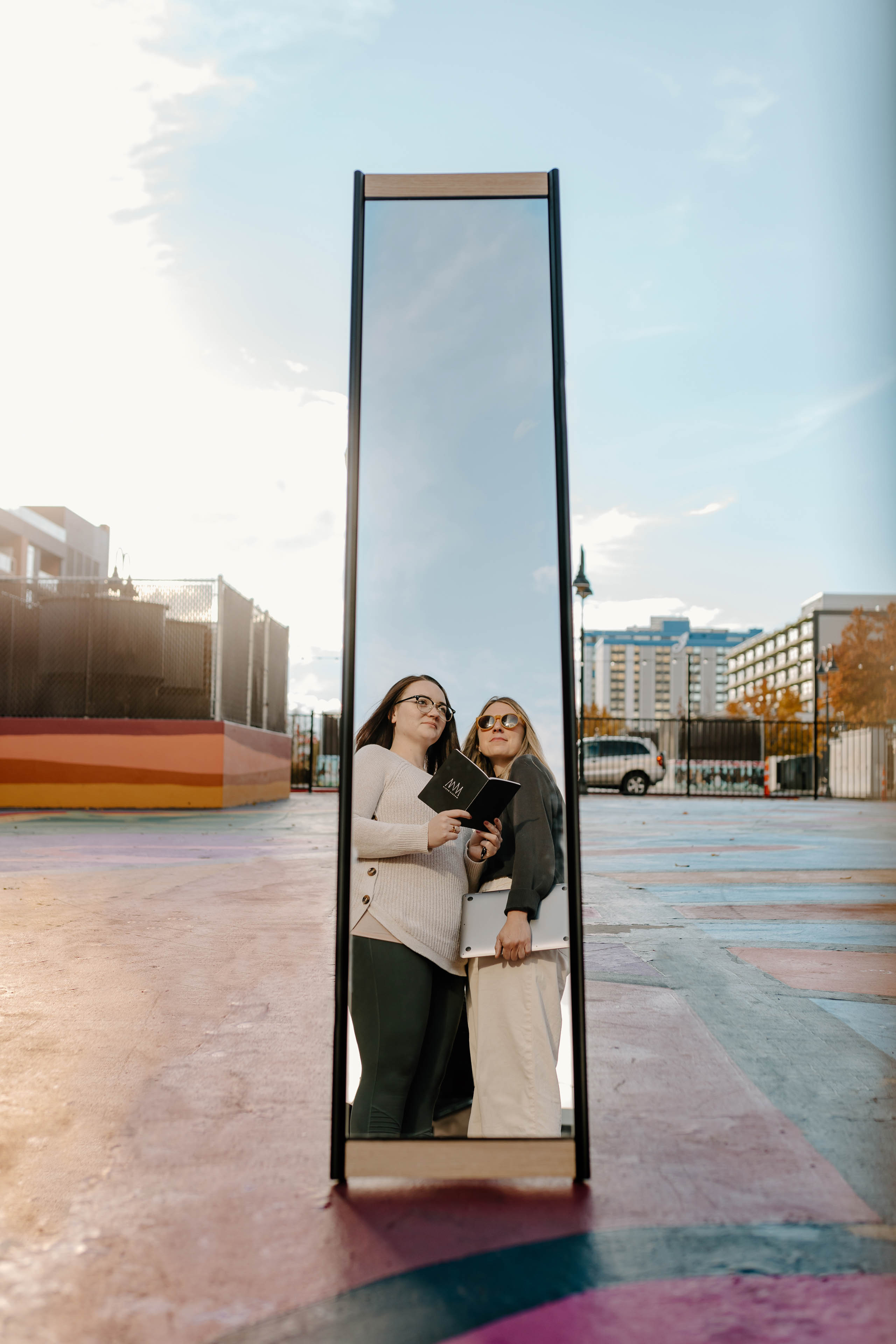 a mirror reflecting two women standing in a city holding a laptop and notebook