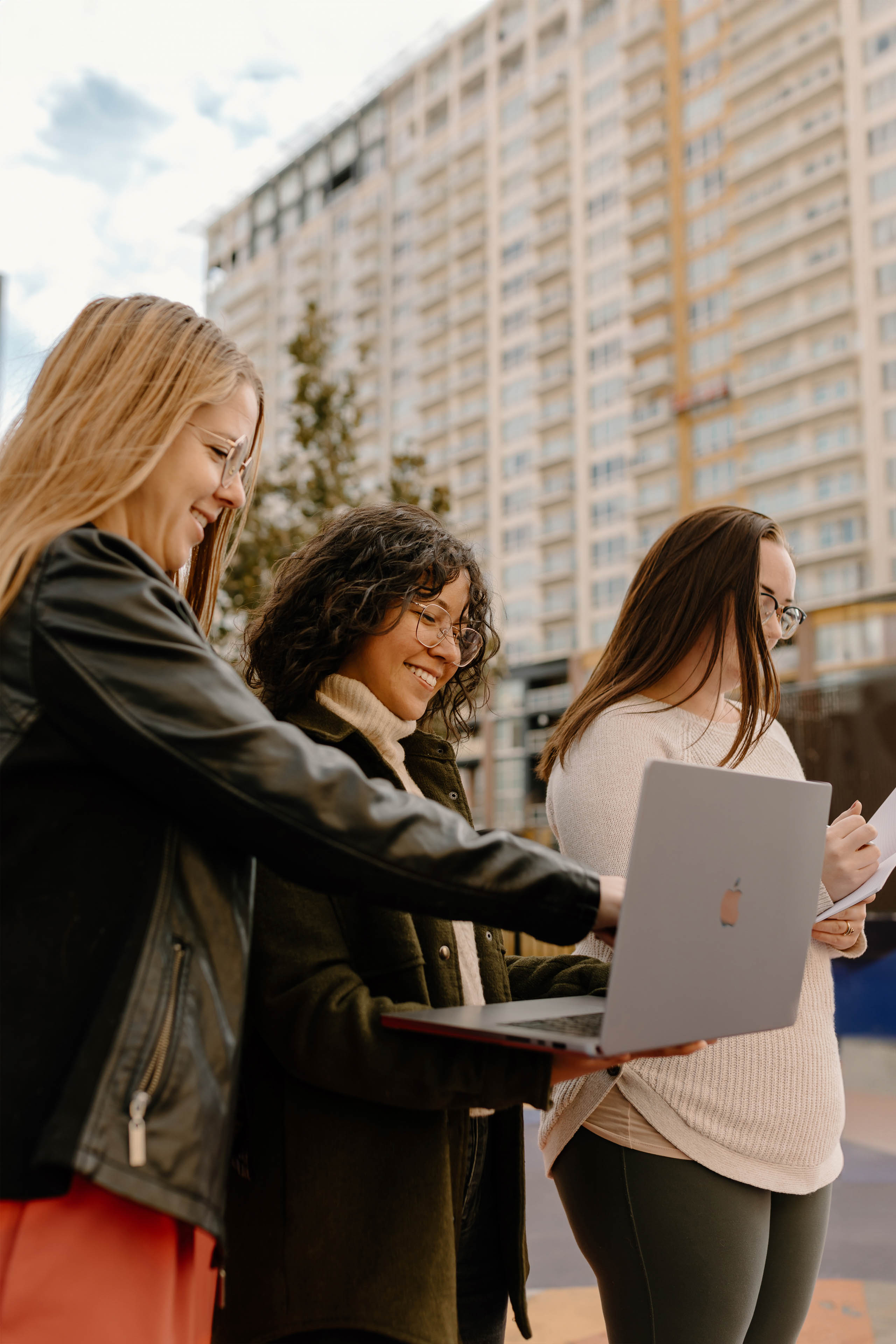 three women standing in a city holding a laptop and notebook