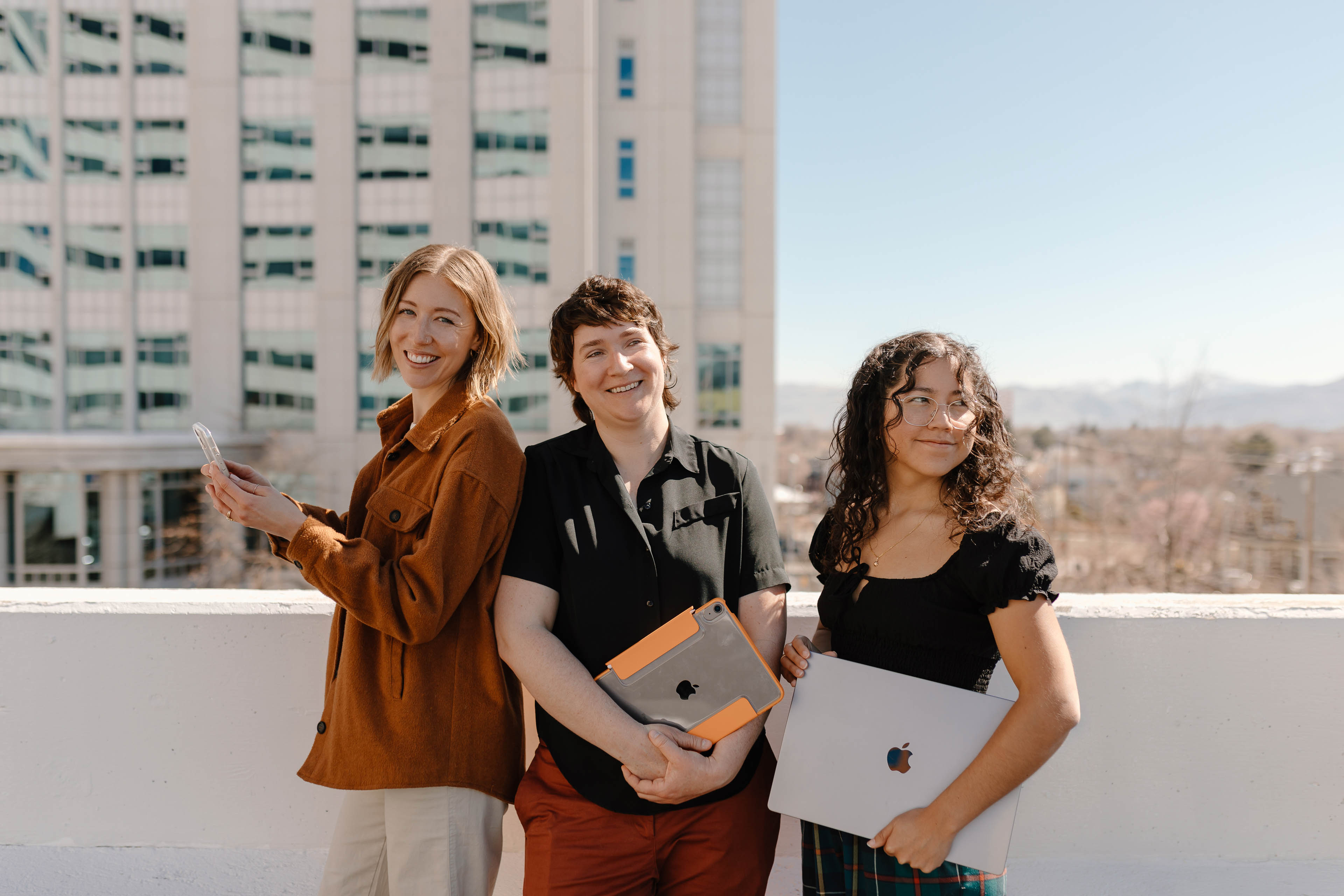 three women standing in a city holding a laptop ipad and iphone